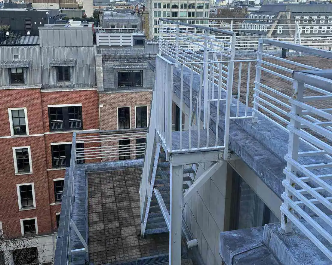 Cluttered rooftop with white-painted escape stairs and railings above neighbouring roofs.